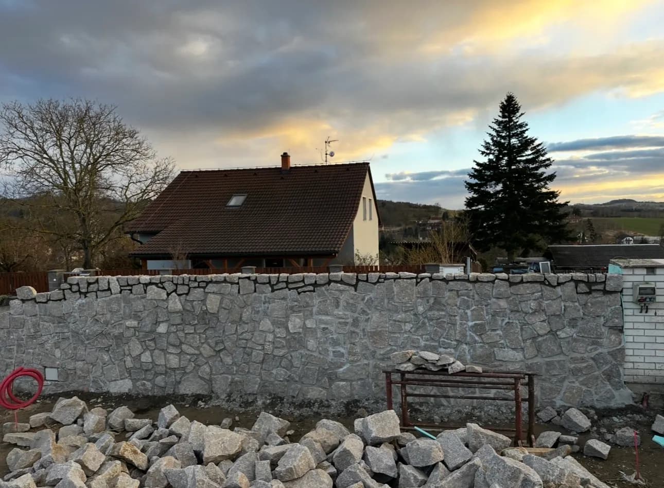 Kamenná plotová zeď dokončená podvečer, v pozadí rodinný dům